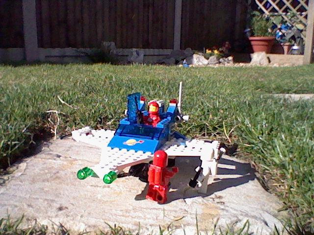 Photo of three Lego astronaut minifigures beside a Lego spaceship. The spaceship is resting on a square stone which is surrounded by grass. Plants and rocks can be seen in the background.