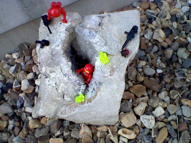 Two red astronauts are exploring a large rock with an open split down the middle. The split is lined with small white crystals. Resting on the sides of the rock are two yellow Lego crystals and some equipment the astronauts brought along with them.