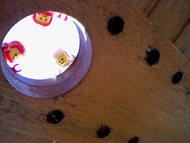 Photo shows the underneath of a wooden garden table. Looking down from a hole in the middle are the three astronauts; their hands hang over the edge of the hole in a way that looks like they are waving to the viewer!