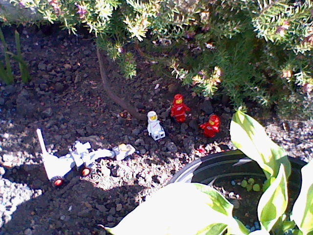 A zoomed out shot of the three astronauts relaxing under the shade of a tall plant. A beam of light passes over a small grey Lego set of a lunar vehicle with a shovel on the end.